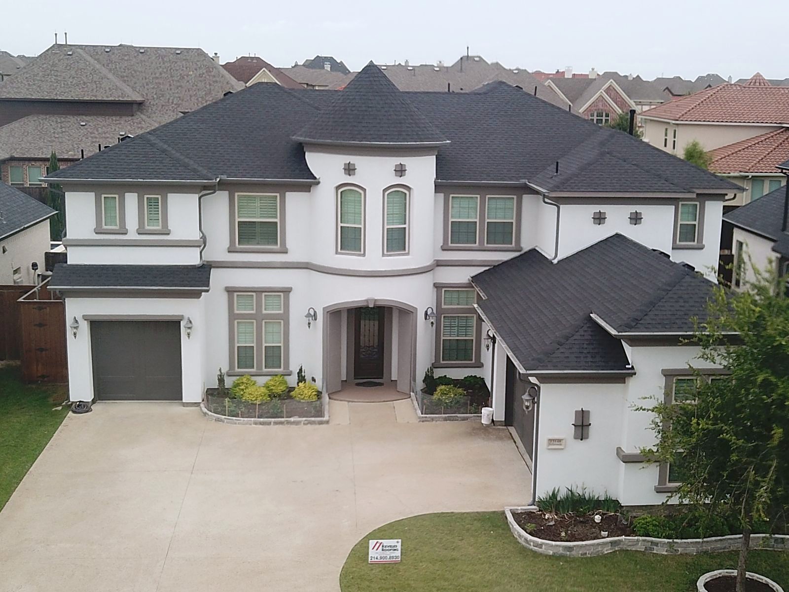 A finished Reveles Roofing job: stone and stucco two-story with a new dark shingle roof, company yard sign on the lawn